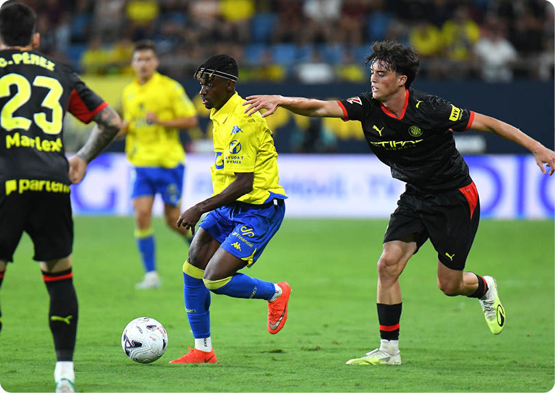 People in sports uniforms are actively playing soccer on a grass field with one person in yellow and blue controlling the ball while others in black and yellow attempt to defend. Descripci n generada con IA