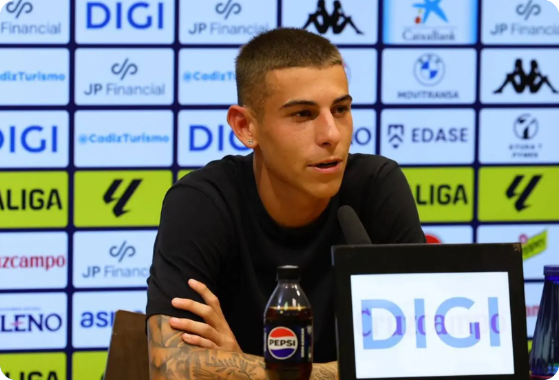 A person wearing a sports jersey is sitting at a press conference table with a microphone and a bottle of Pepsi in front of a backdrop covered with various sponsor logos. Descripci n generada con IA