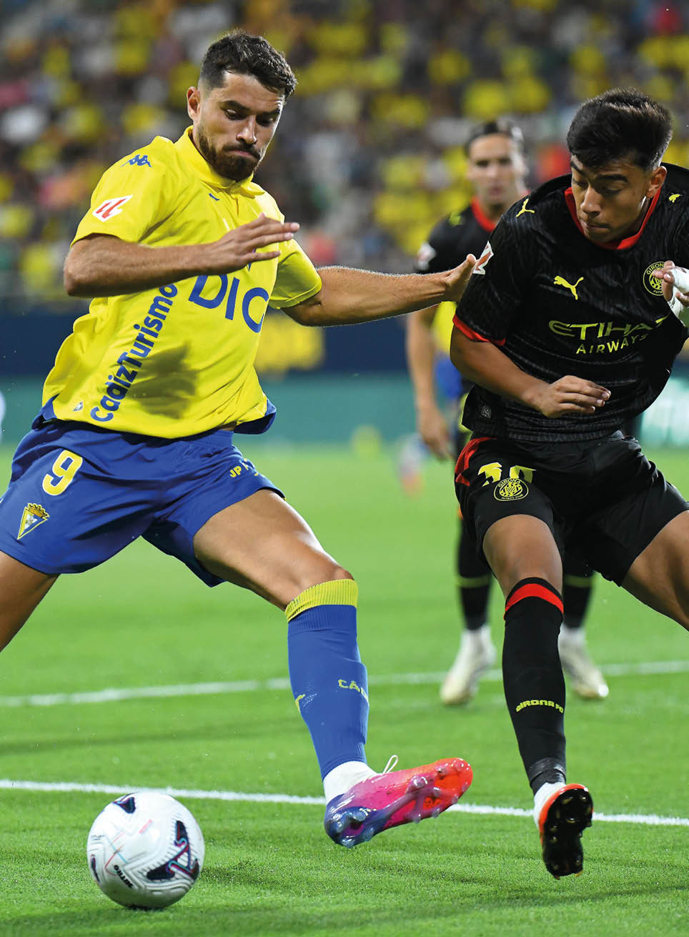 A person wearing a yellow and blue soccer uniform is kicking a soccer ball on a grassy field while another person in a black uniform runs nearby. Descripci n generada con IA