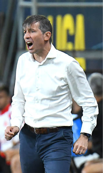 A person wearing a white shirt and dark pants is walking or moving with a focused expression in an indoor setting with blurred people and yellow signage in the background. Descripci n generada con IA