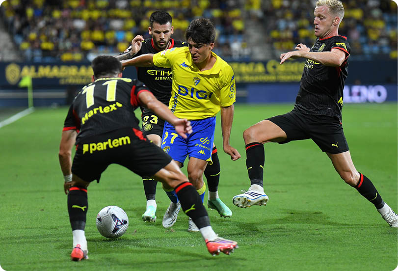 Four people in sports uniforms are actively competing for a soccer ball on a grass field in a stadium. Descripci n generada con IA
