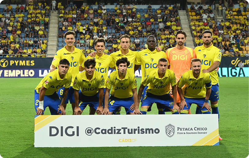 A group of adults wearing yellow and blue sports uniforms pose together on a soccer field behind a sign promoting Cadiz tourism and a seafood export company with a stadium crowd in the background. Descripci n generada con IA