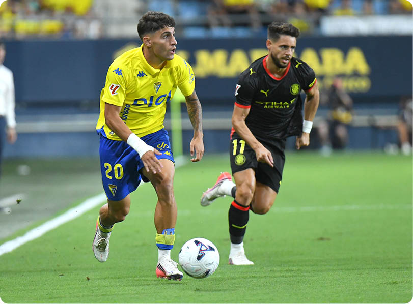 Two adults in sports uniforms are actively running on a soccer field with one controlling the ball while the other chases closely behind. Descripci n generada con IA