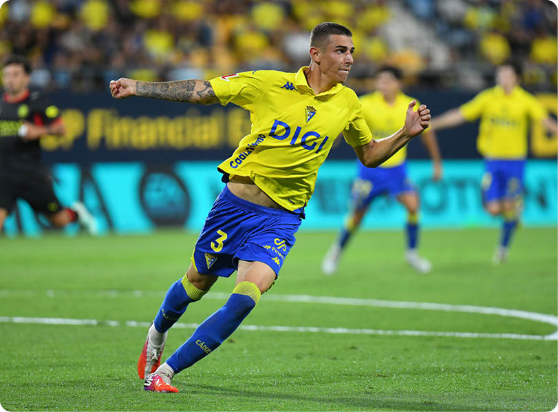 A person in a yellow and blue sports uniform is running on a soccer field during a game with other players and a referee visible in the background. Descripci n generada con IA