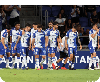 Jugadores de f tbol con uniforme azul y blanco caminando juntos en un campo durante un partido con p blico en las gradas. Descripci n generada con IA