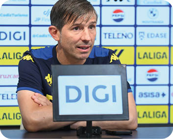 Una persona con camiseta deportiva azul y amarilla est sentada con los brazos cruzados frente a un micr fono en una conferencia de prensa con un fondo lleno de logotipos.  Descripci n generada con IA