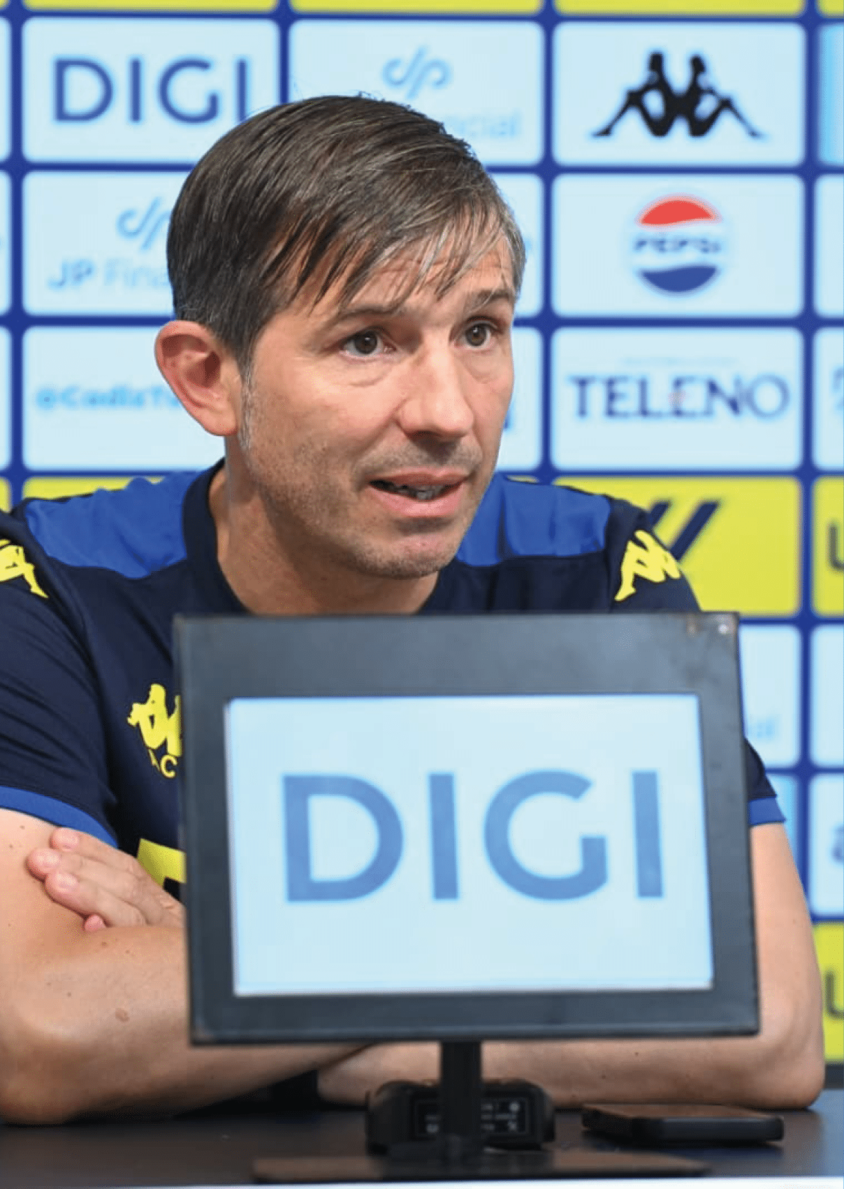 Una persona con camiseta deportiva azul y detalles amarillos est sentada frente a un micr fono con un cartel que dice DIGI en un entorno que parece una conferencia de prensa deportiva.  Descripci n generada con IA
