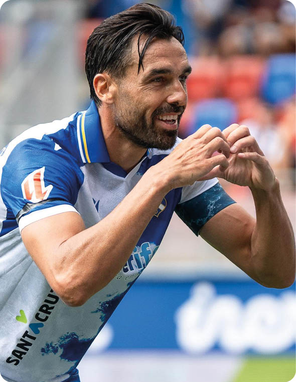 Una persona con camiseta deportiva blanca y azul sonr e mientras forma un coraz n con las manos en un entorno de estadio. Descripci n generada con IA