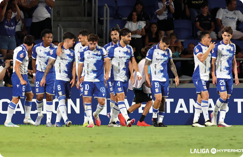 Un grupo de personas con uniformes deportivos blancos y azules camina sobre un campo de c sped en un estadio con p blico al fondo. Descripci n generada con IA