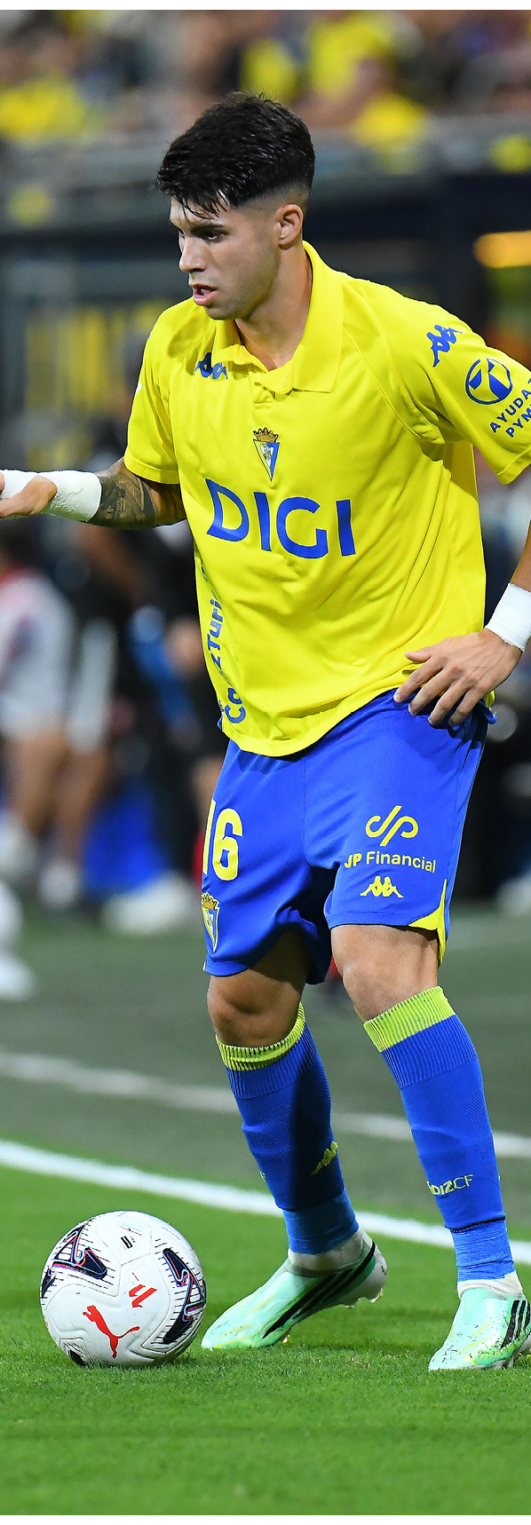 Persona con uniforme deportivo amarillo y azul controlando un bal n de f tbol en un campo. Descripci n generada con IA