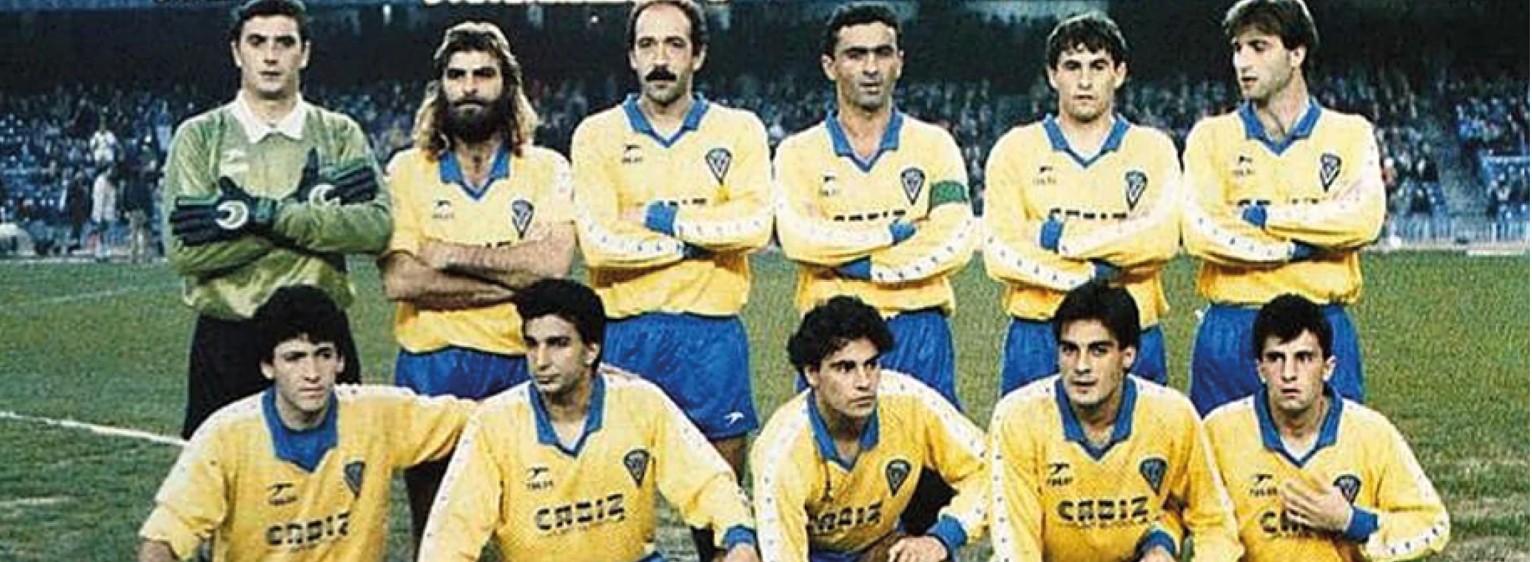 Un grupo de adultos con uniformes deportivos amarillos y azules posan en un campo de f tbol con un estadio y espectadores al fondo. Descripci n generada con IA