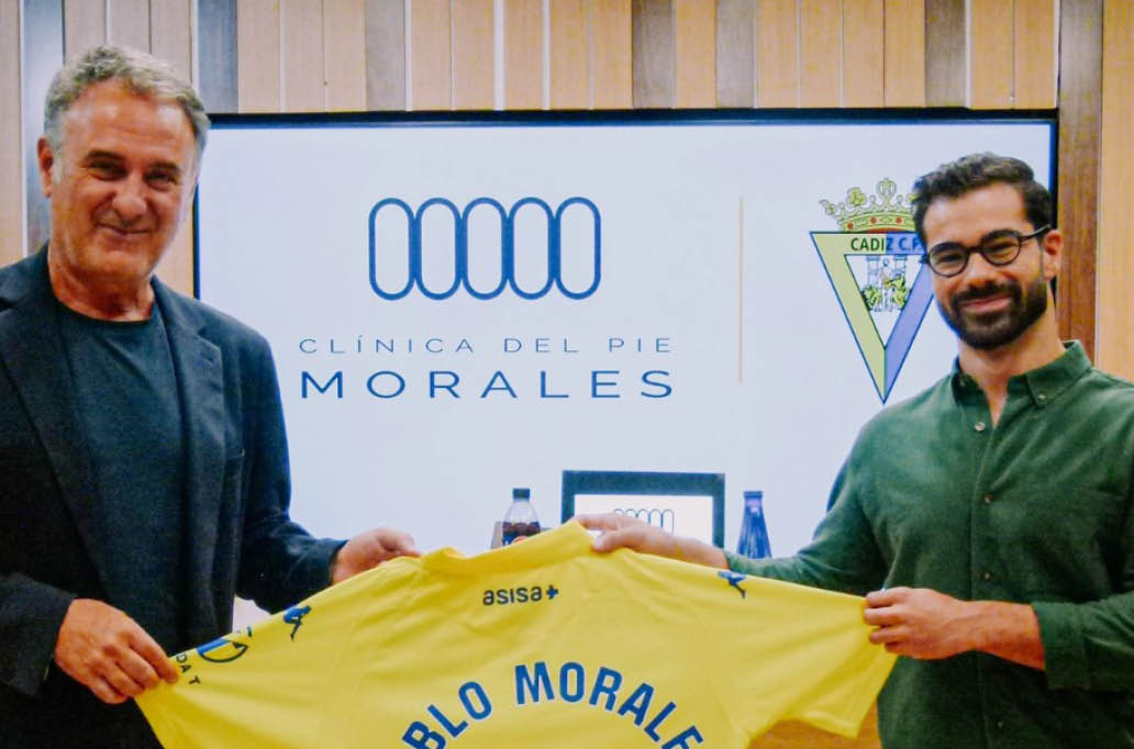 Dos personas sonrientes sostienen una camiseta amarilla con el texto “BLO MORALE" frente a un cartel que dice "Cl nica del Pie Morales". Descripci n generada con IA