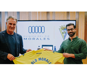 Dos personas sonrientes sostienen una camiseta amarilla frente a un cartel que dice Cl nica del Pie Morales en un ambiente interior con paredes de madera. Descripci n generada con IA