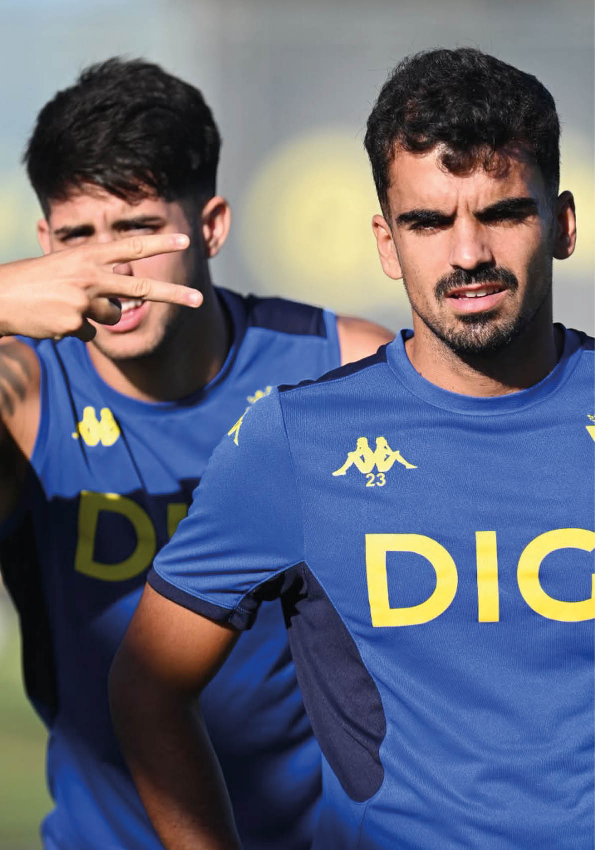 Dos personas con camisetas deportivas azules con el logo DIGI est n al aire libre, una al frente con expresi n seria y la otra detr s haciendo un gesto con la mano. Descripci n generada con IA