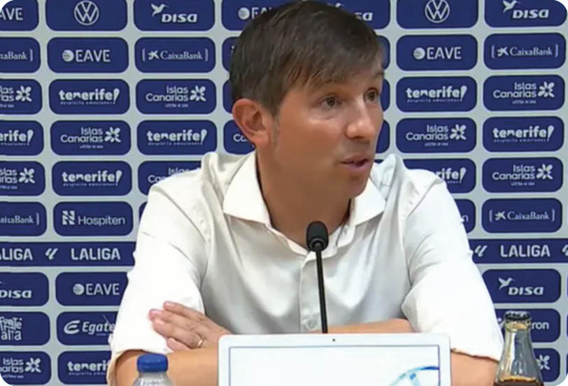 A person with short hair wearing a white shirt is speaking into a microphone at a press conference with a backdrop covered in logos. Descripci n generada con IA