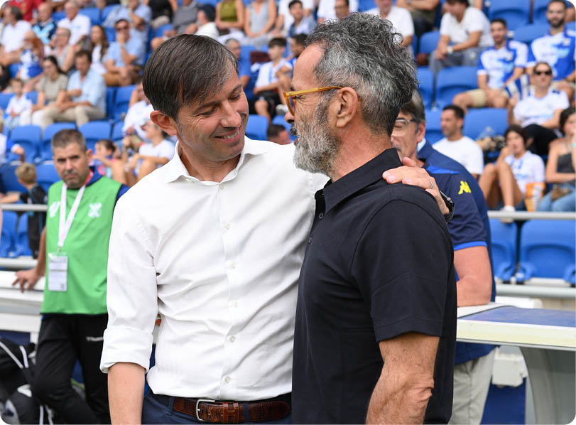Two adults are smiling and embracing each other in a stadium with seated spectators in the background. Descripci n generada con IA