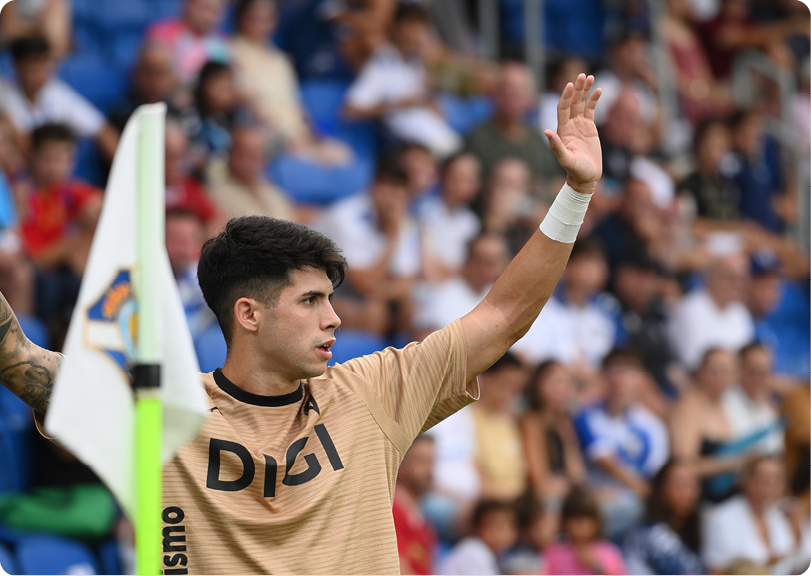 A person wearing a beige sports jersey with the word DIGI raises one arm while standing near a corner flag on a sports field with a blurred crowd in the background. Descripci n generada con IA
