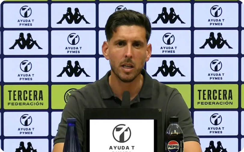 A person with short dark hair wearing a dark polo shirt is speaking at a press conference with a backdrop featuring logos and text. Descripci n generada con IA