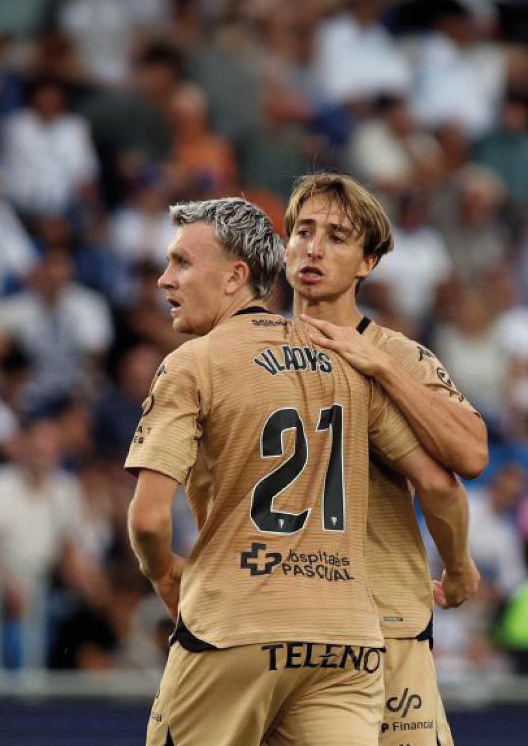 Two adults wearing matching sports jerseys with the number 21 are embracing on a field with a blurred crowd in the background. Descripci n generada con IA