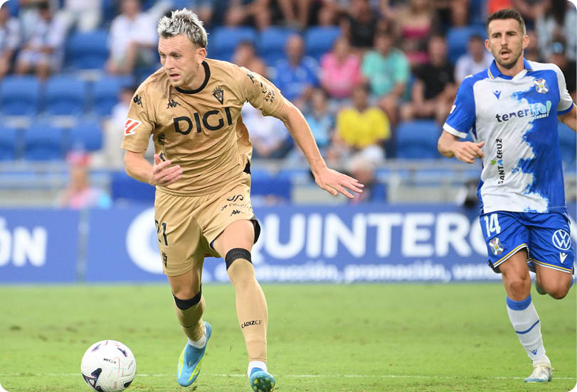 Two adults in soccer uniforms are actively running on a soccer field with one person closely following the other who is controlling the ball. Descripci n generada con IA