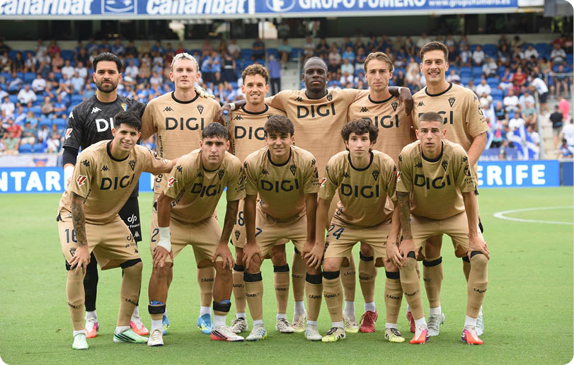 A group of eleven adults wearing matching gold sports uniforms with the word DIGI on the front are posing together on a soccer field with a stadium crowd in the background. Descripci n generada con IA