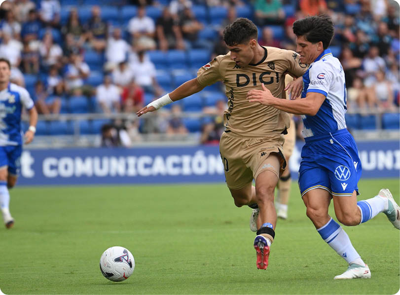 Two people in sports uniforms are competing for a soccer ball on a grassy field with spectators in the background. Descripci n generada con IA