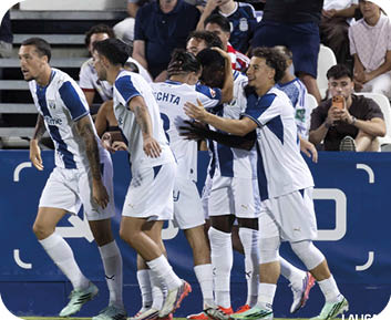 Un grupo de personas con uniformes deportivos blancos y azules celebran juntos en un campo de f tbol mientras algunas personas observan desde las gradas. Descripci n generada con IA
