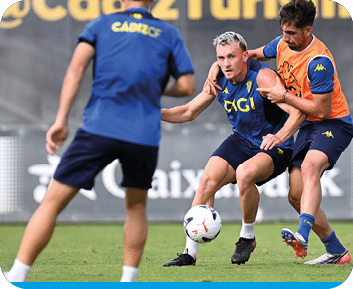 Tres personas con ropa deportiva est n en un campo de f tbol, una de ellas controla el bal n mientras otra intenta quitarle la pelota y la tercera observa. Descripci n generada con IA