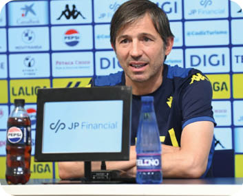Una persona con camiseta deportiva azul con detalles amarillos est sentada en una mesa frente a un micr fono y dos botellas, con un fondo lleno de logotipos de patrocinadores.  Descripci n generada con IA