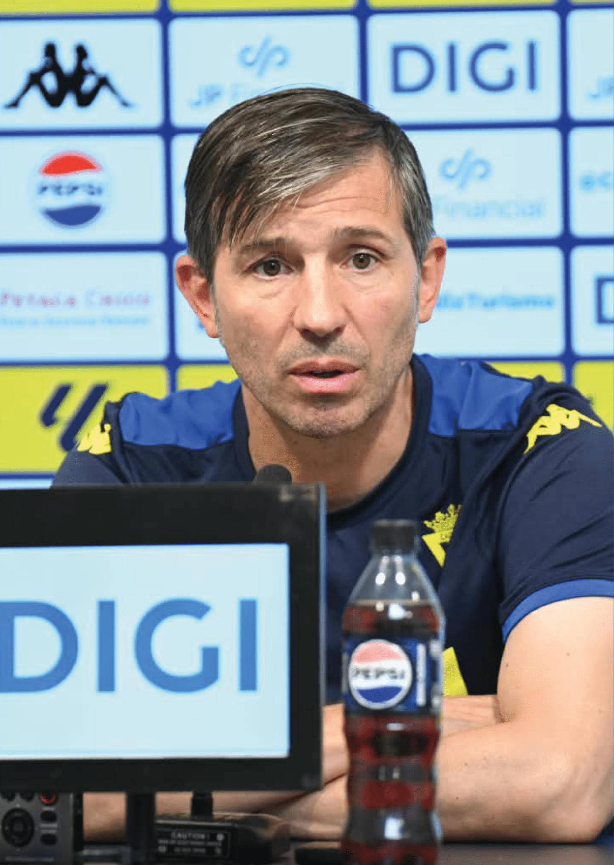 Una persona con camiseta deportiva azul con detalles amarillos est sentada frente a un micr fono y una botella de bebida en una conferencia de prensa con un fondo de logotipos.  Descripci n generada con IA