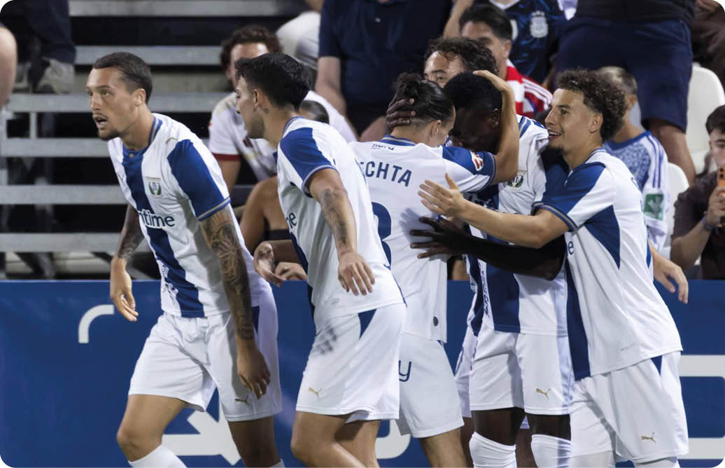 Un grupo de personas con uniformes deportivos blancos y azules celebran en un campo de f tbol mientras otras personas observan desde las gradas. Descripci n generada con IA