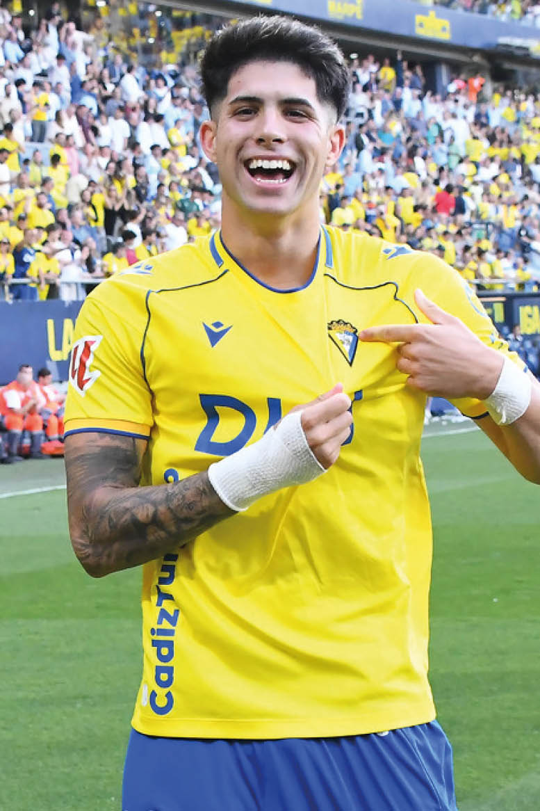 Una persona sonriente con camiseta amarilla y pantalones azules est en un campo de f tbol con p blico animado al fondo.  Descripci n generada con IA
