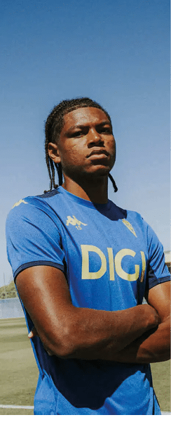 Persona con camiseta azul con el logo de DIGI posando con los brazos cruzados en un campo de f tbol bajo un cielo despejado. Descripci n generada con IA