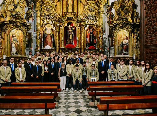 Grupo grande de personas vestidas formalmente y con uniformes posando en el interior de una iglesia decorada con elaborados retablos dorados y figuras religiosas. Descripci n generada con IA