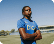 Una persona con trenzas lleva una camiseta deportiva azul con el logo DIGI y est de pie con los brazos cruzados en un campo de f tbol bajo un cielo despejado.  Descripci n generada con IA