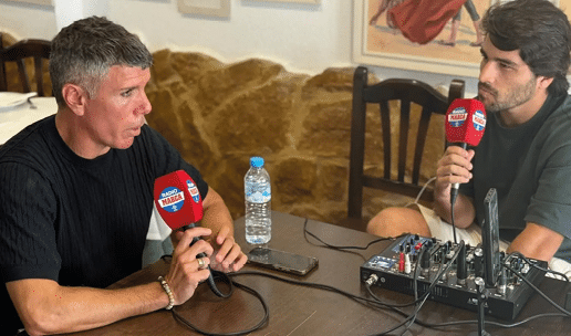 Dos personas sentadas frente a frente en una mesa sosteniendo micr fonos rojos mientras participan en una entrevista o conversaci n grabada en un ambiente interior con decoraci n en la pared. Descripci n generada con IA