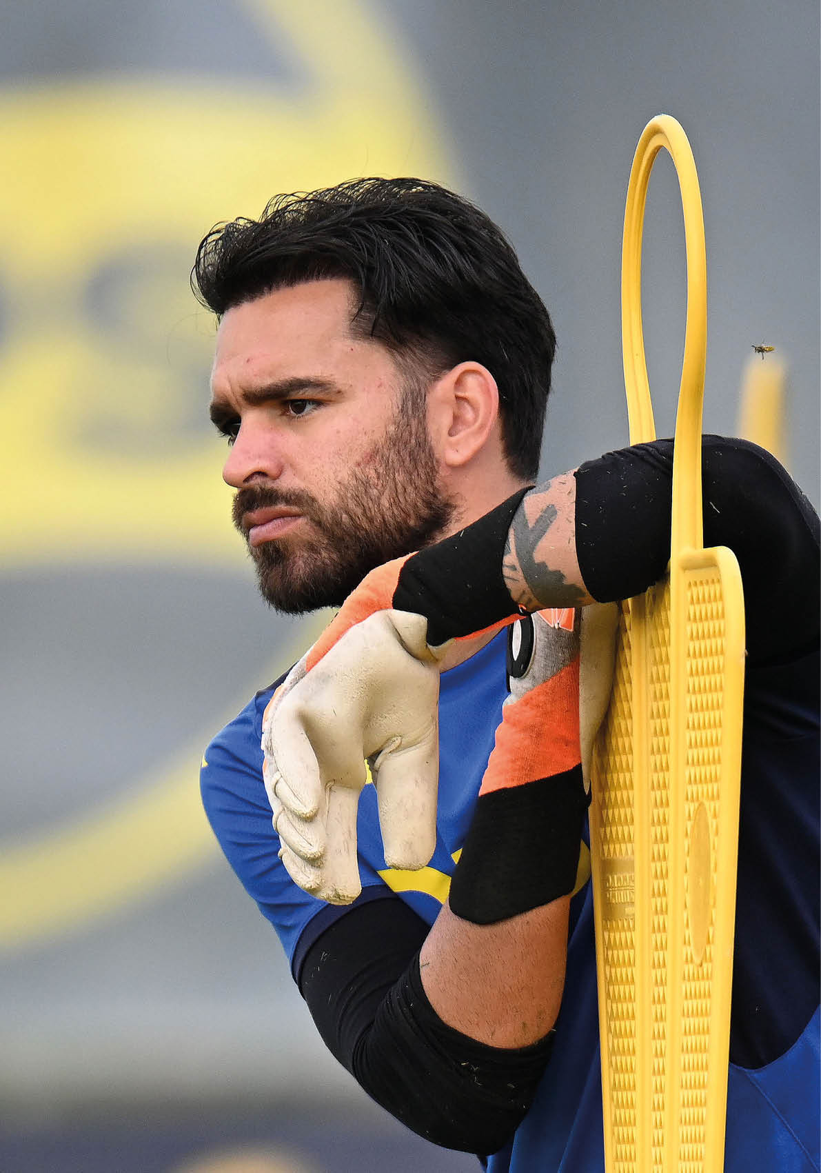 Una persona con guantes de portero y camiseta azul se apoya pensativa en un accesorio de entrenamiento amarillo en un campo deportivo. Descripci n generada con IA