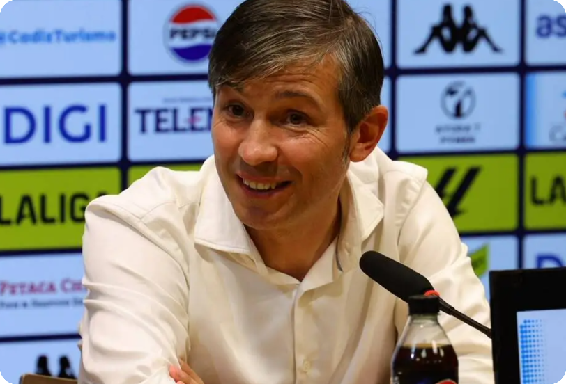 A person with short hair wearing a white shirt is speaking into a microphone at a press conference with a backdrop covered in logos. Descripci n generada con IA