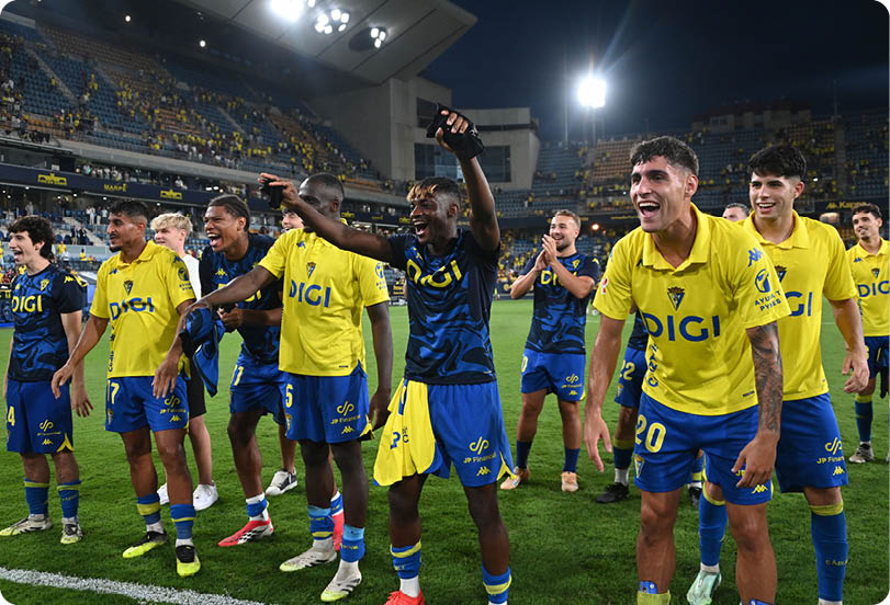 A group of adults wearing yellow and blue sports uniforms are celebrating energetically on a soccer field in a stadium at night. Descripci n generada con IA
