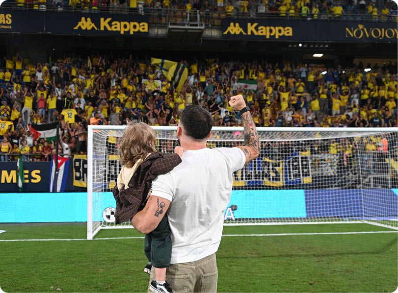 A person holding a child is raising a fist in front of a soccer goal with a large crowd of fans wearing yellow in the stadium stands. Descripci n generada con IA