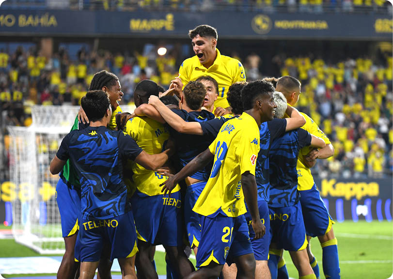 A group of people wearing yellow and blue sports uniforms are celebrating together on a soccer field with one person lifted above the others. Descripci n generada con IA