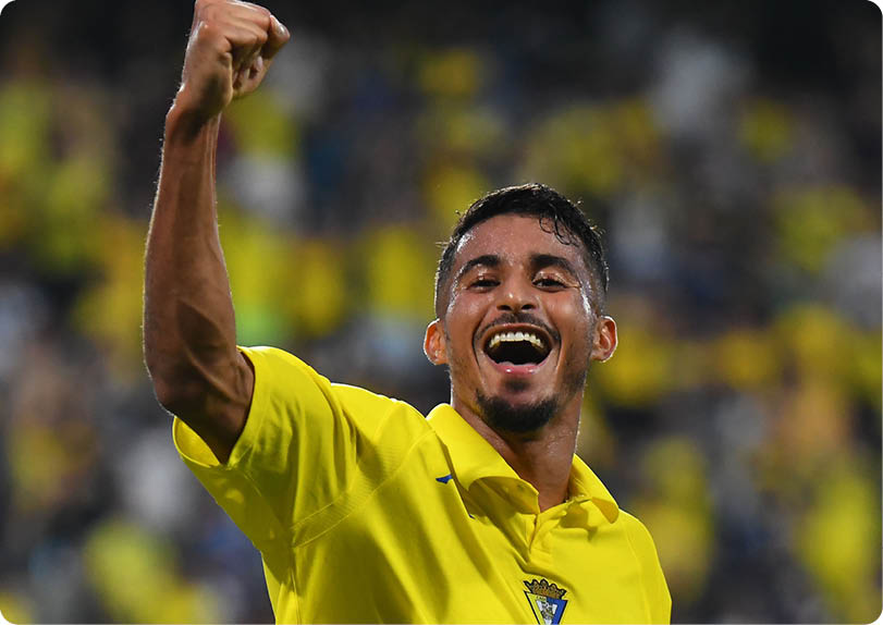 A person wearing a yellow shirt is celebrating with a raised fist and a joyful expression in a blurred stadium setting. Descripci n generada con IA