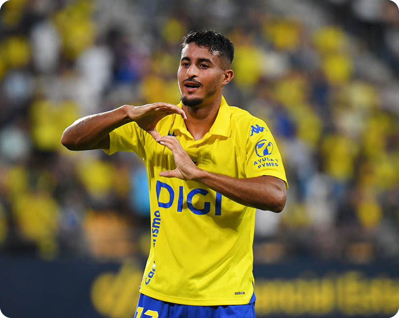 A person wearing a yellow sports jersey and blue shorts is making a heart shape with their hands on a sports field with a blurred crowd in the background. Descripci n generada con IA