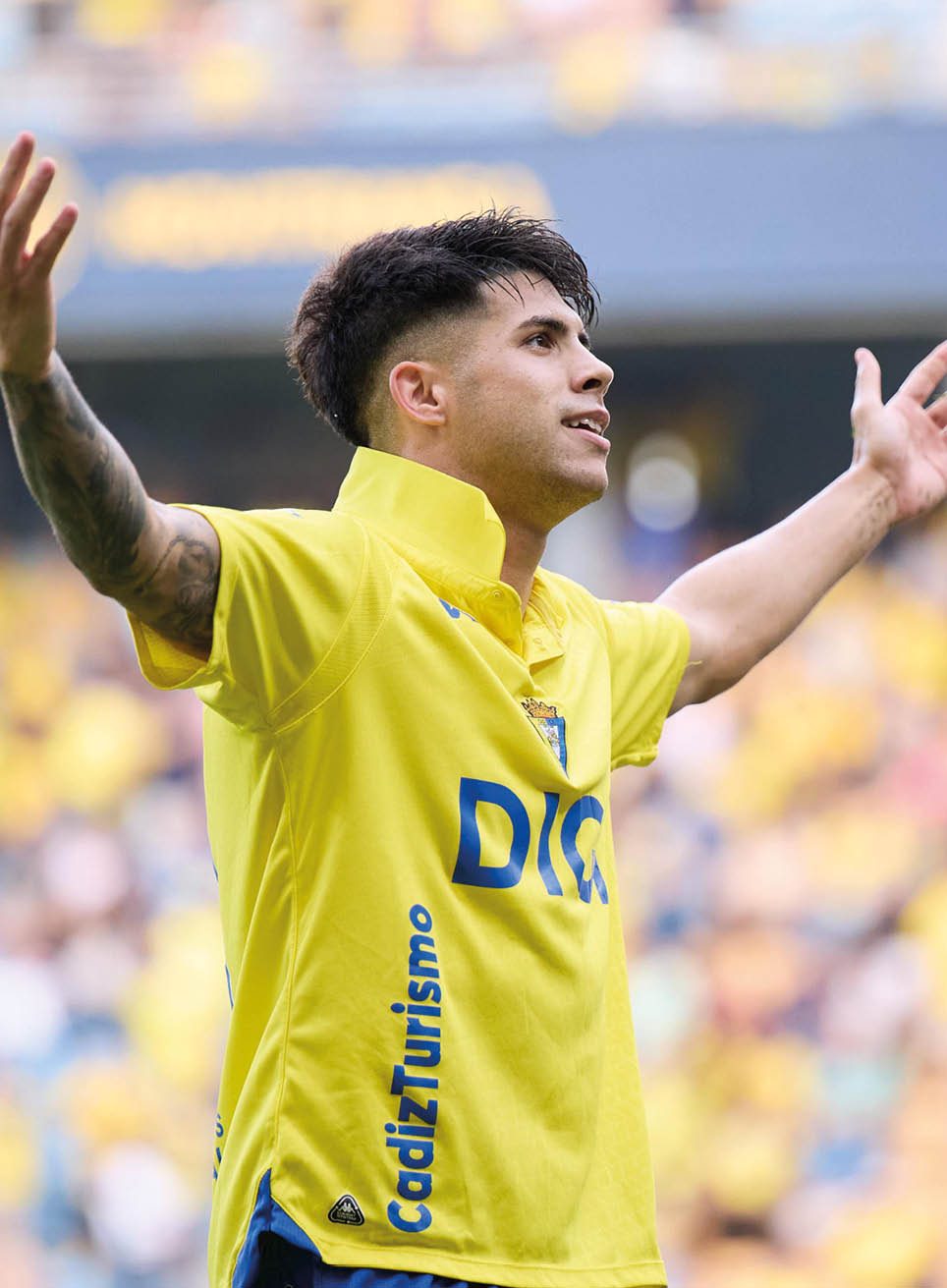 A person with short dark hair wearing a yellow sports jersey is raising their arms and looking to the side with a focused expression. Descripci n generada con IA