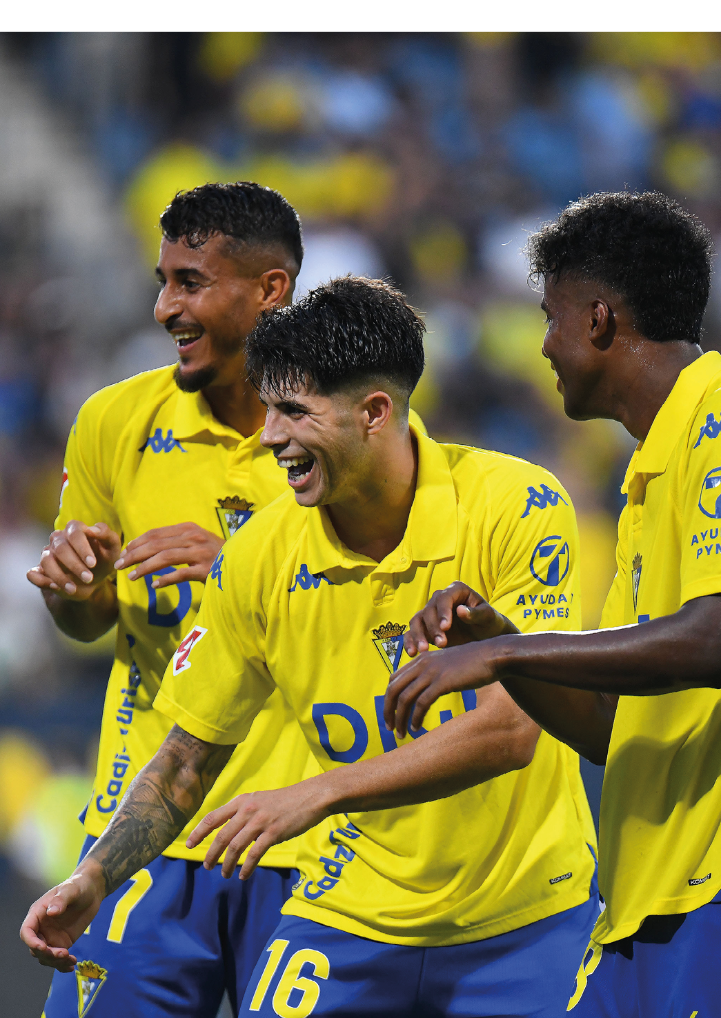Three people wearing yellow and blue sports uniforms are smiling and interacting energetically on a sports field. Descripci n generada con IA