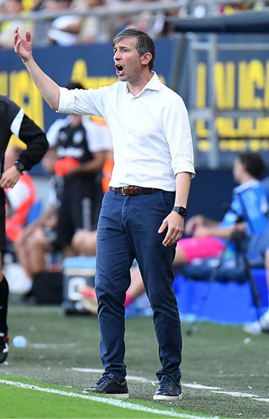 A person in a black sports outfit stands on the sideline of a soccer field while another person in a white shirt and blue pants gestures animatedly. Descripci n generada con IA