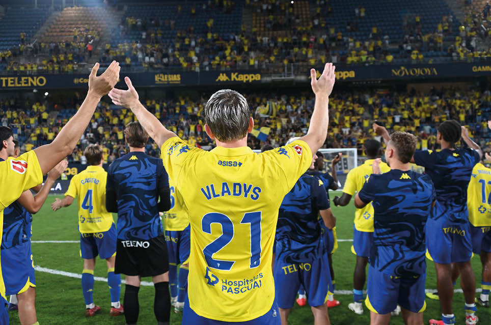 People wearing yellow and blue sports uniforms are standing on a soccer field with their arms raised, facing a stadium crowd. Descripci n generada con IA