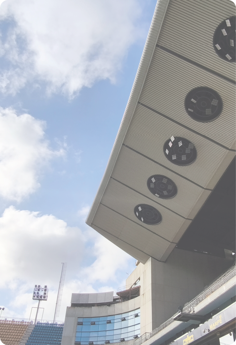 A stadium roof with large circular speakers or lights is shown against a partly cloudy sky above a section of seating and a building with reflective windows. Descripci n generada con IA
