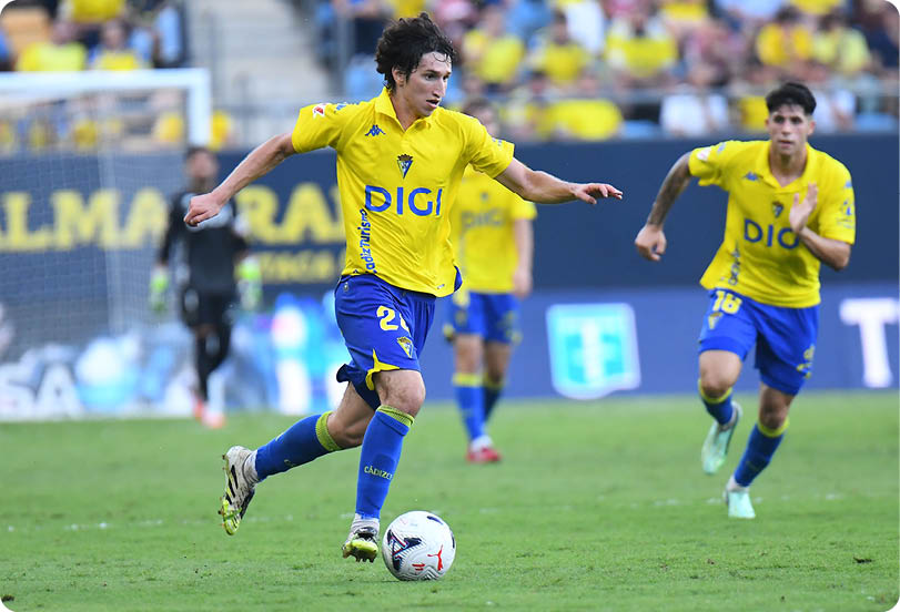 Two adults in yellow and blue soccer uniforms are actively playing on a grass field with one controlling the ball and the other running nearby. Descripci n generada con IA