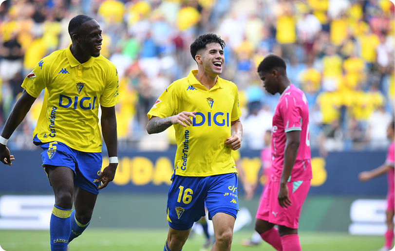 Two people in yellow and blue soccer uniforms are celebrating while a person in a pink uniform walks nearby on a soccer field with a blurred crowd in the background. Descripci n generada con IA